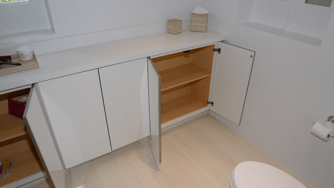 Bathroom vanity run with doors open showing the internal carcass, adjustable shelves and concealed hinges beneath a seamless solid-surface worktop
